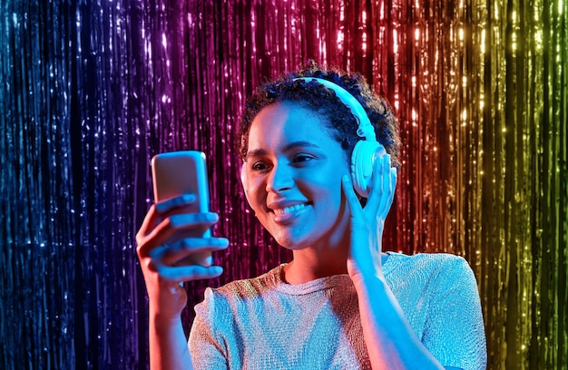 People at the party wear wireless headphones with different color lights while selecting music on their phone, surrounded by a festive decor with colourful lights.
