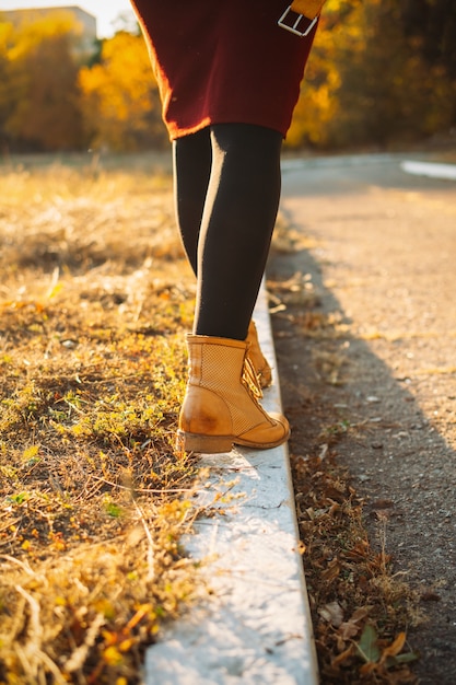 A close-up of a woman's legs as she walks on a paved path with autumn leaves falling around her. She is wearing stylish athletic shoes and the focus is on the movement and the seasonal setting of the walk.