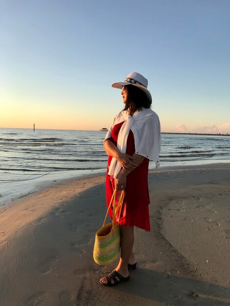 A woman in her 60s wearing a white linen shirt, cream-colored pants, and a straw hat, walking along a sandy beach at sunset. She is holding a woven tote bag and smiling serenely.