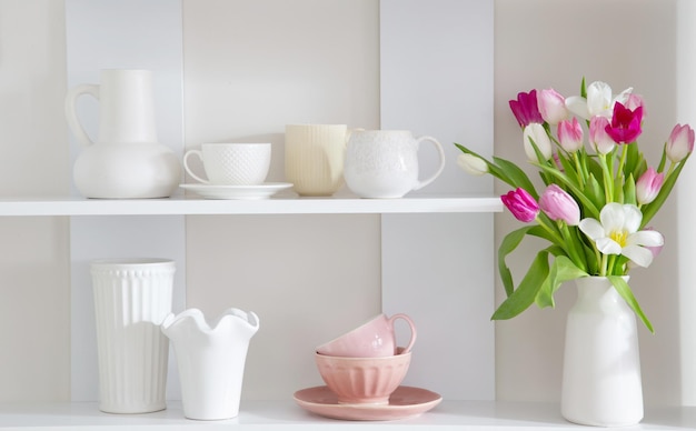 A brightly lit kitchen with white cabinets, marble countertops, and open shelving displaying white ceramic dishes and glassware. A large window overlooks a lush garden.