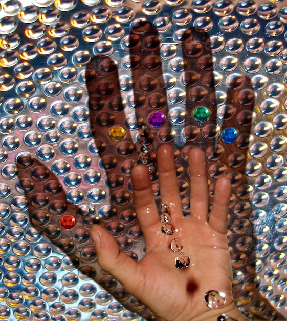 A hand showcasing maximalist nail art with a mix of glitter, tiny rhinestones, and 3D floral charms on a bright, multicolored background. The background is blurred, putting the emphasis on the intricate details of the nails.