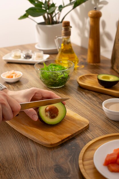 A brightly lit tabletop featuring a bowl of homemade face mask, a cut avocado halves, a lemon, and a jar of honey, with a woman's hand stirring the mask in the background.