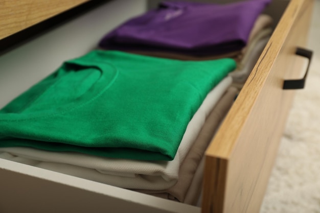 A close-up shot of a well-organized drawer containing neatly folded neutral-colored sweaters and t-shirts. Labels identify the contents of each section, emphasizing the organization and simplicity of a curated wardrobe. The drawer is part of a minimalist, light wood dresser.