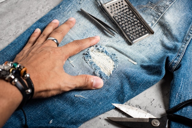 A person cutting denim jeans to create shorts, showcasing the DIY process of repurposing old clothes. The image captures the hands in action, with scissors and denim scraps visible on the table.